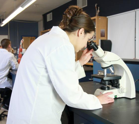 Nursing student in lab coat looking through microscope in lab
